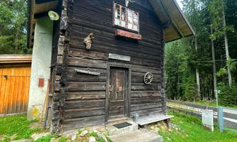 A rustic wooden house in a quiet forest setting. The building has a simple architecture and is surrounded by green areas.