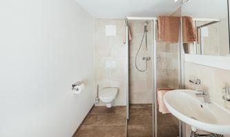 A modern bathroom with a shower cabin, a sink, and a toilet. The walls are bright and the floor is tiled.