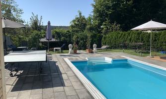 A beautiful garden with a blue swimming pool and a table tennis table. The sun is shining and there are umbrellas for shade.