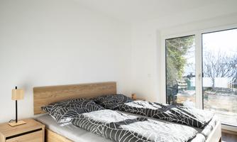 A modern bedroom with a wooden bed and bright bedding. There is a nightstand with a lamp and plenty of daylight through the large window.