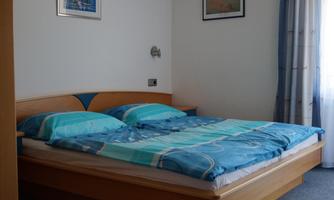 A cozy bedroom with a large double bed and blue bedding. The walls are light, and there are windows with curtains.