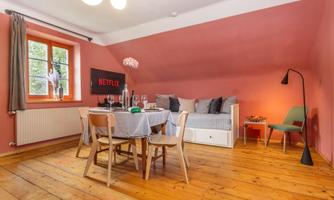 A cozy living space with a dining table, chairs, and a couch. The walls are painted in a warm pink, and a large window provides plenty of light.