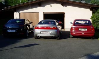Three cars are parked in front of a garage in a quiet environment. The sun is shining and there is plenty of greenery nearby.