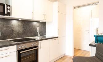 A modern kitchen with white cabinets and a black countertop. In the background, a bright room and an open door are visible.