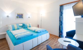 A modern bedroom with a large bed and bright blue bedspreads. The room has plenty of light and a comfortable seating area.