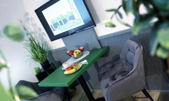 A modern room with a green table and comfortable gray chairs. On the table is a fruit bowl, and in the background is a television with the logo "CITY".
