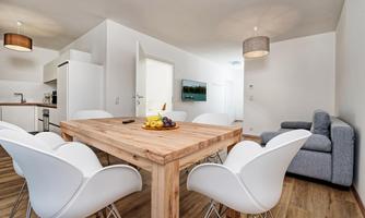 A modern dining room with a large wooden table and white chairs. In the background, a kitchen and a cozy sofa can be seen.