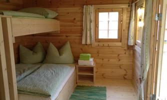 A cozy sleeping room made of wood with a bunk bed and a seating area. The walls are light and wooden, with green cushions and a rug on the floor.