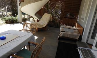 A terrace with a table and chairs made of rattan. In the background, there is a lounge chair and a wooden privacy screen.