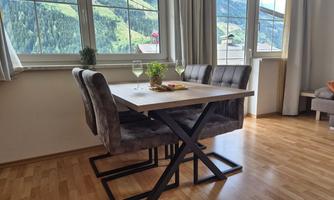 A modern dining table with four chairs is in a cozy room. Large windows offer a view of the green mountains.