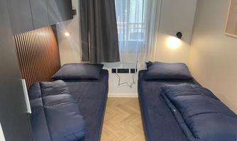 A cozy bedroom with two single beds and modern furnishings. The room has bright walls and daylight streaming through a window.