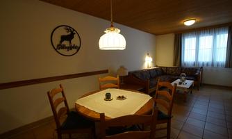 A cozy interior with a round table and chairs. In the background, there is a sofa and a window with curtains.