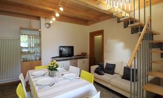 A bright, cozy living and dining room with a table for six people. A wooden staircase leads to the upper floor, and there is a modern couch with cushions.