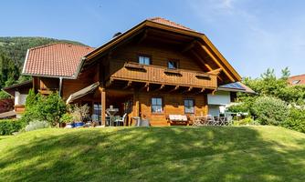 A beautiful wooden house with a balcony and a well-maintained garden. In the background, green mountains and a blue sky can be seen.