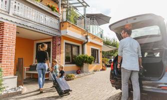 A woman with a suitcase is walking with two children to a cozy home. A man is standing next to a car and unloading luggage.