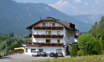 A charming building in the mountains with colorful flowers on the balconies. In the foreground, cars are parked and the surroundings are lush and scenic.