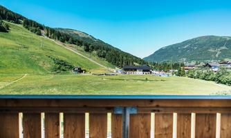 A beautiful view of green meadows and hills under a bright blue sky. In the background, some buildings and a mountainous landscape can be seen.