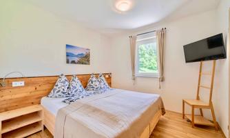 A modern bedroom with a double bed and wooden furniture. There is a television on the wall and plenty of daylight coming through the window.