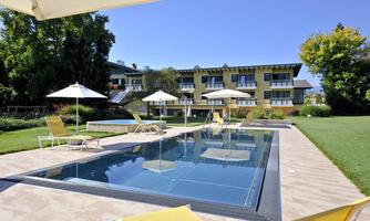 A beautiful pool area with loungers and sun umbrellas in a well-maintained garden. In the background, there is a modern building.