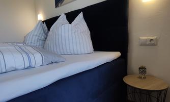 A cozy bedroom with a blue bed and pillows. A picture of a beautiful landscape hangs on the wall.