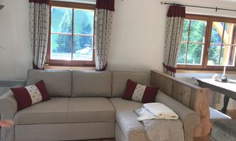 A cozy living room with a beige sofa and red cushions. Large windows offer a view of nature.
