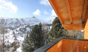 A picturesque view of snow-covered mountains and forests from a balcony. The sky is clear and the sun is shining.