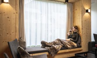 A cozy interior view with a woman sitting relaxed by the window enjoying a glass of wine. Soft blankets and wooden paneling create a warm atmosphere.