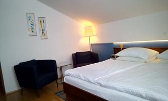 A cozy bedroom with a large bed and two chairs. The walls are painted light and there is a soft light from a floor lamp.