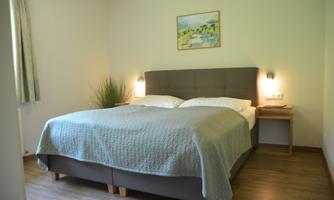 A bright bedroom with a cozy double bed and a small bedside table. A picture hangs on the wall, and the windows let in soft light.