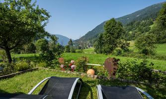 A beautiful garden with lounge chairs, surrounded by lush greenery and mountains in the background. The clear sky and nature create a relaxed atmosphere.