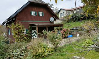 A charming wooden house with a green garden. In the background, more houses and trees can be seen.