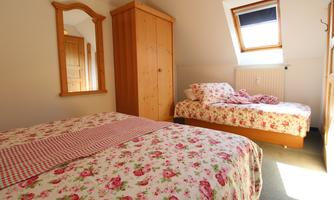 A cozy bedroom with two beds and a flower pattern. There is a wooden wardrobe and a window with daylight.