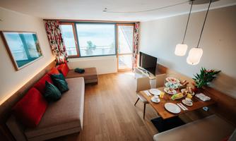A modern living room with a cozy sofa and a dining table. Large windows provide a view of the landscape.