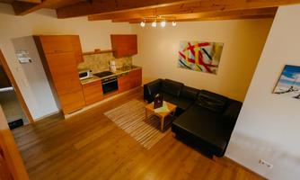 A modern living room with a black couch and a small table. In the background, there is an open kitchen with wooden cabinets and a colorful painting on the wall.