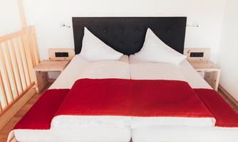 A modern bedroom with a double bed and white pillows. The duvet is red and the room has a bright, friendly atmosphere.