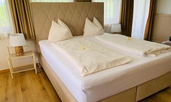 A bright bedroom with a large double bed and two pillows. Next to the bed are two nightstands with lamps.