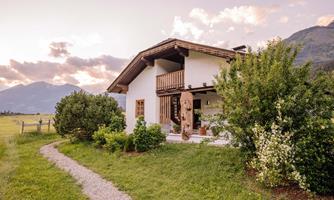 A charming house in a green meadow with mountains in the background. The path to the house is lined with trees and bushes.