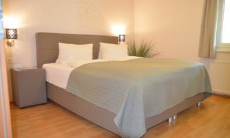 A cozy bedroom with a large bed and pretty bedding. The walls are light and there are decorative elements like bedside lamps and plants.