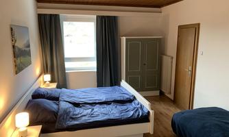 A cozy bedroom with two beds and blue bedding. Next to the beds are nightstands with lamps and a dresser in the background.