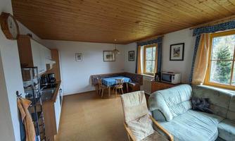 A bright living room with wooden floor and ceiling. There is a cozy seating area, a table, and a kitchenette.