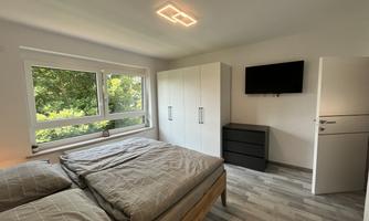 A modern bedroom with a large bed and bright walls. The room has large windows, a television, and provides a view of greenery.
