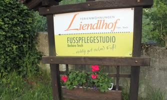 A wooden sign with the inscription "Liendlhof" and "Foot Care Studio" indicates where the holiday apartments are located. Various flowers adorn the lower shelf of the sign.