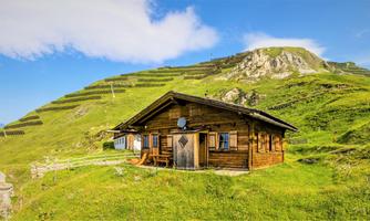 A cozy wooden holiday cabin stands on a green meadow. In the background, gentle hills and a blue sky are visible.
