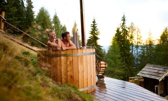 A couple relaxes in a wooden bathtub in nature. In the background, there are trees and a picturesque landscape.