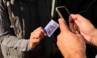 A person hands a card to another person while holding a smartphone in their hand. The scene appears to take place outdoors.