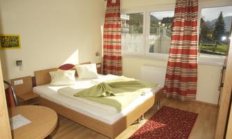 A simple bedroom with two single beds, a table, and chairs. Large windows provide ample daylight and a cozy atmosphere.