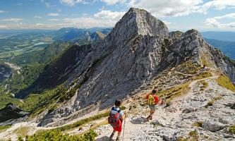 Two hikers explore a rocky mountain path with stunning views of the surrounding valleys and mountains. The sky is blue and cloudy.
