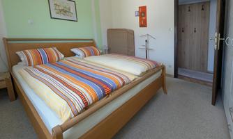 A cozy bedroom with a large double bed and colorful striped bedding. In the background, there is a wardrobe and an open room with a door.