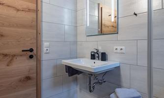 A modern bathroom with a wooden cabinet door and a simple sink. The walls are light tiled, and there is a large mirror.