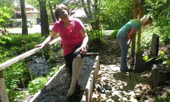 Two women are standing on a narrow wooden walkway in a green forest. One woman is sitting with one leg in the water, while the other is busy in the background.